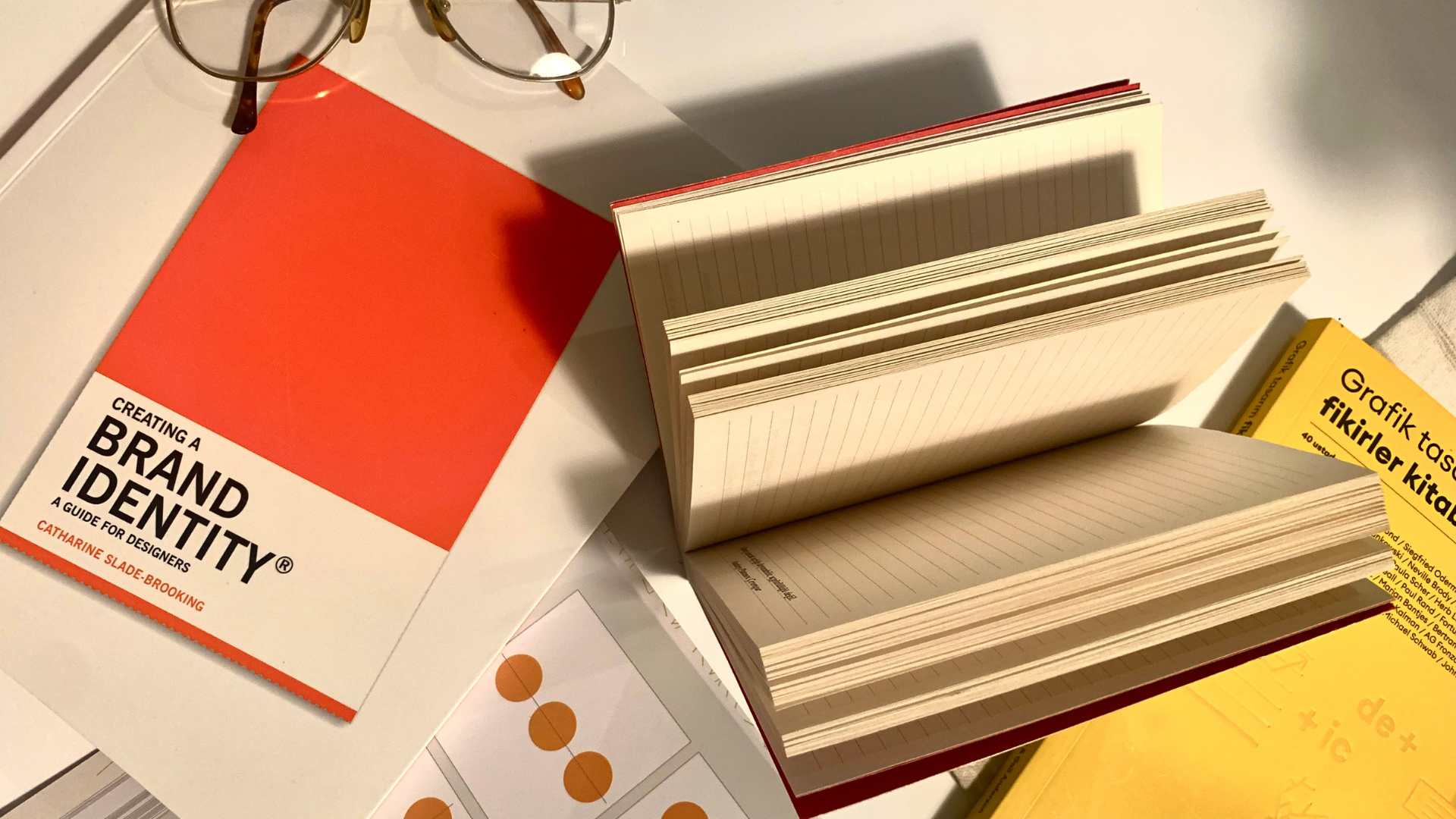 Brand identity design books and a lined notebook open on a desk beside a pair of glasses
